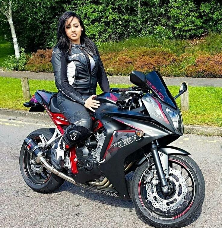A person in a black leather jacket and protective gear sits on a black and red sport motorcycle parked on a road, with green bushes and trees in the background.