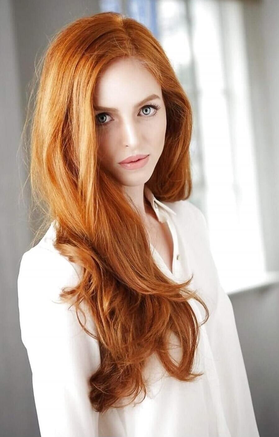 Portrait of a woman with long, wavy red hair, wearing a white blouse.