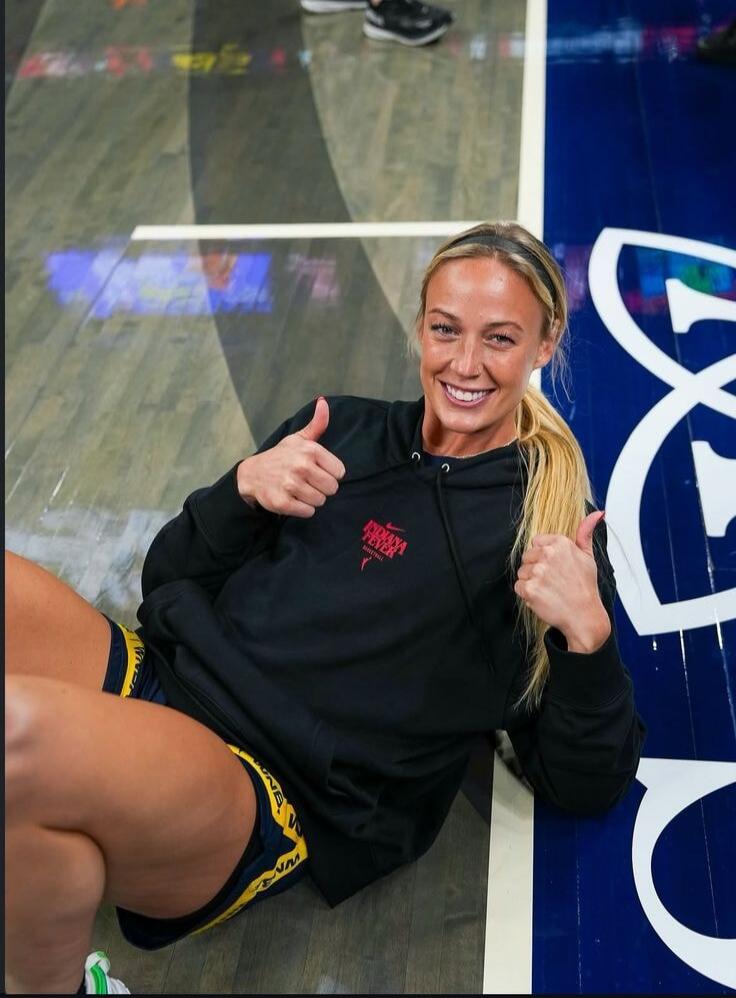 INDIANA FEVER BASKETBALL. WNBA. A woman is sitting on a basketball court, smiling and giving two thumbs up.