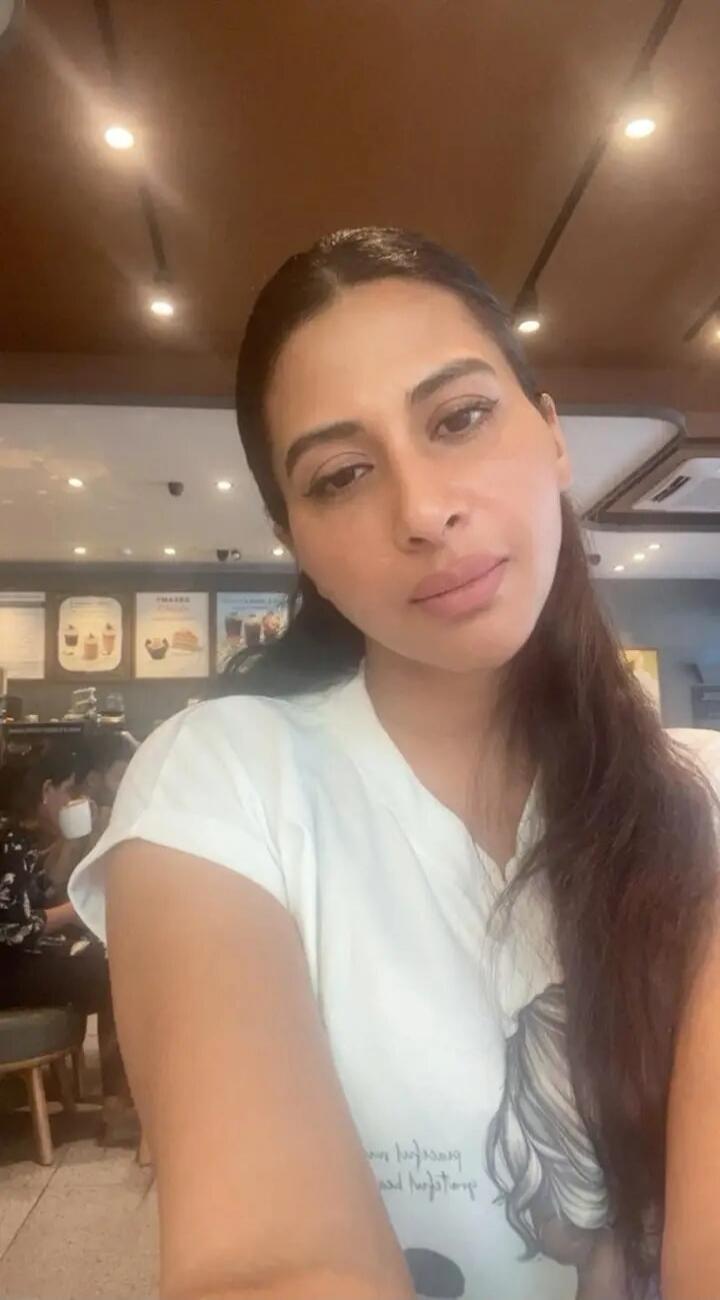 A woman is taking a selfie in a cafe. There are other patrons and cafe decor in the background. Text on her t-shirt is partially visible but unclear.