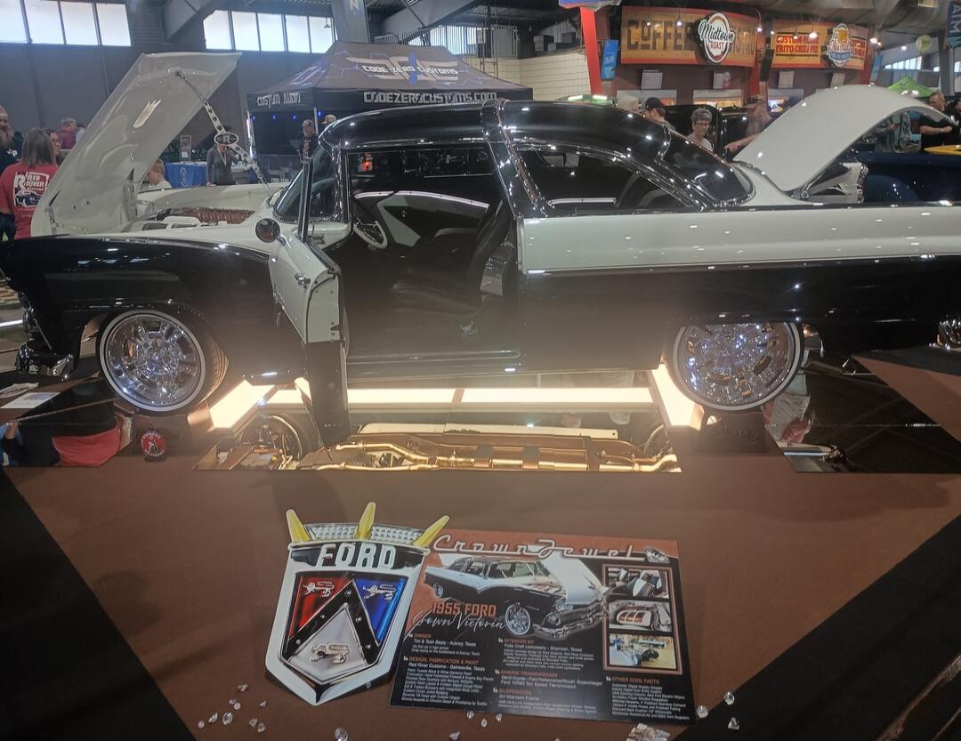 FORD logo displayed on a stand at a car show. A black and white customized classic car is on a raised platform with its doors open and hood up. A display board and promotional cards are in front of the car.