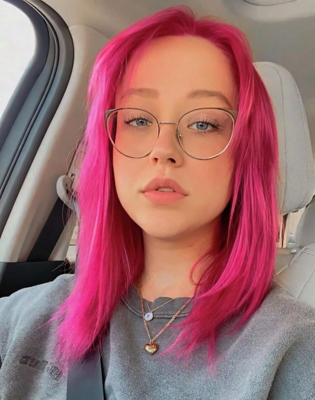 Portrait of a young woman with bright pink hair and glasses sitting in a car.