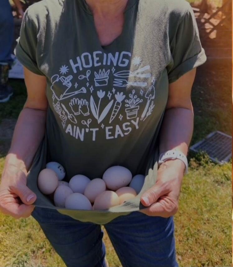 HOEING AIN'T EASY. A person is holding a collection of fresh eggs in the hem of their t-shirt. The t-shirt has a humorous gardening slogan and various gardening tool icons.