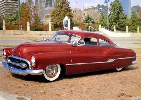 A vintage red car with whitewall tires is parked on a paved surface. In the background, there are modern buildings and trees under a clear sky.