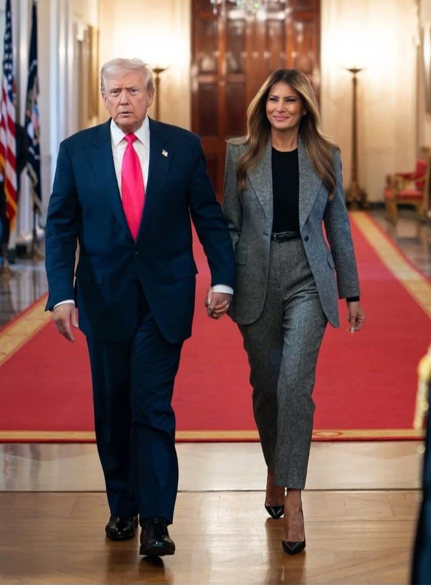Donald Trump and Melania Trump walking hand in hand down a hall with a red carpet.