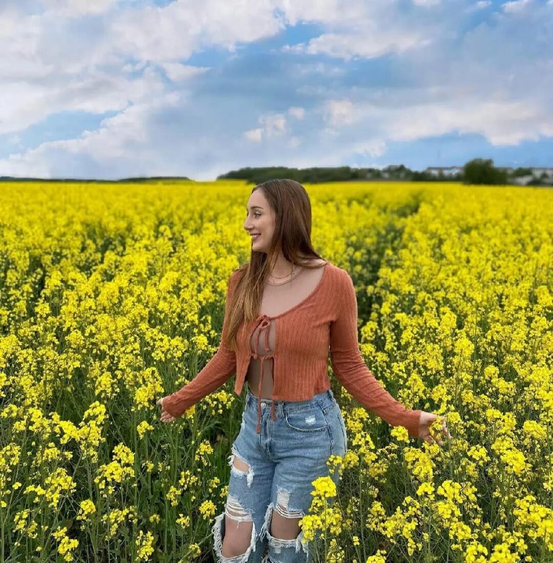 A young woman with long brown hair, wearing a rust-colored tie-front crop top and ripped jeans, stands smiling in a vast field of bright yellow flowers under a blue sky with clouds.
