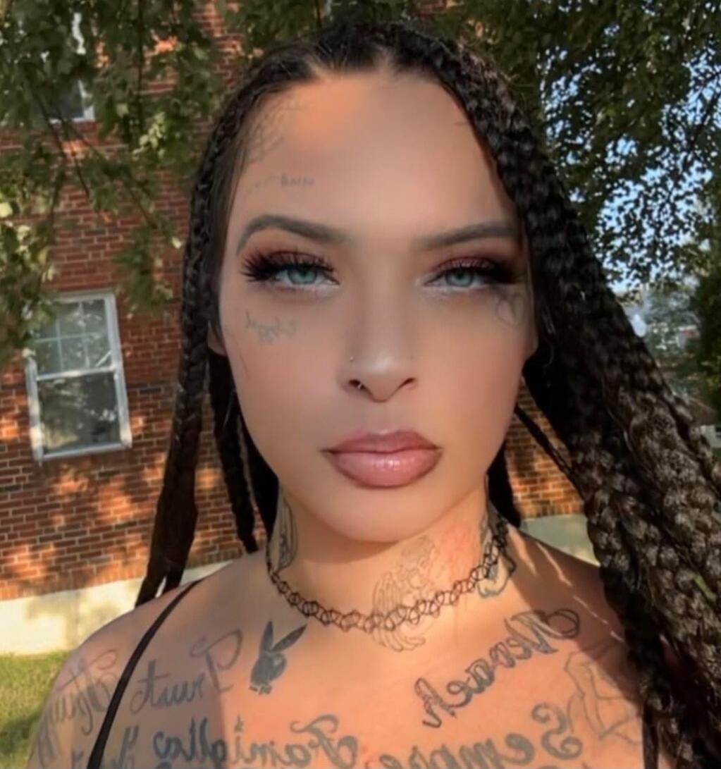 Portrait of a woman with braided hair, visible tattoos on face, neck, and chest, wearing makeup.