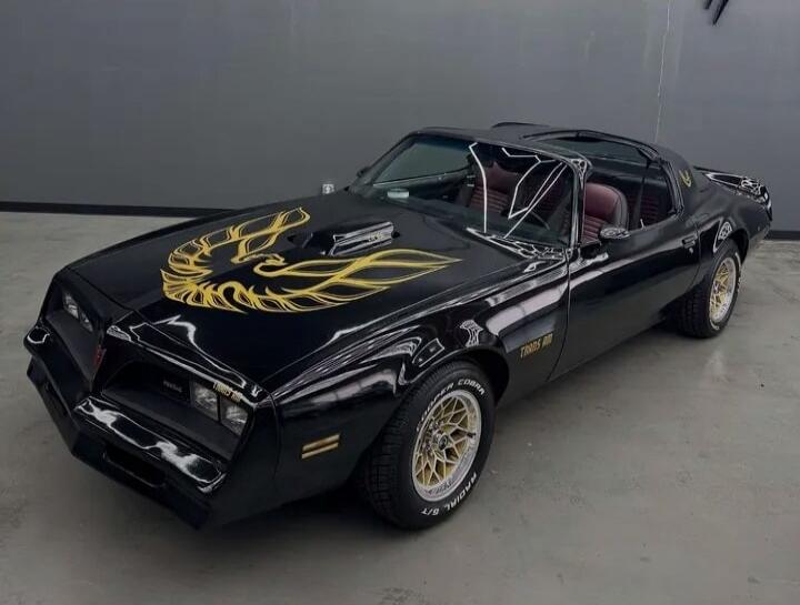 Black classic convertible car featuring a large gold phoenix decal on the hood. The vehicle has gold mesh-style wheels and a tinted interior. The windshield is shattered, giving the impression of damage or a damaged top. Overall, a striking, vintage muscle car appearance.