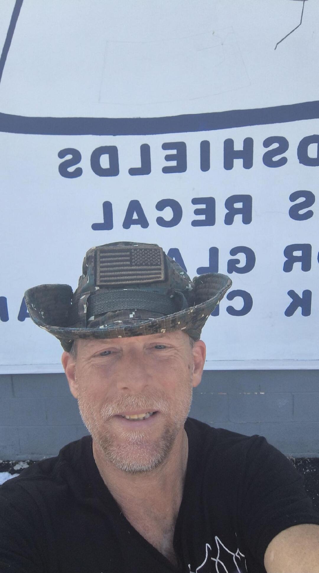 A man wearing a camouflage boonie hat with an American flag patch, smiling. In the background, there is a white wall with blue, mirrored text, which appears to include 'SHIELDS'.