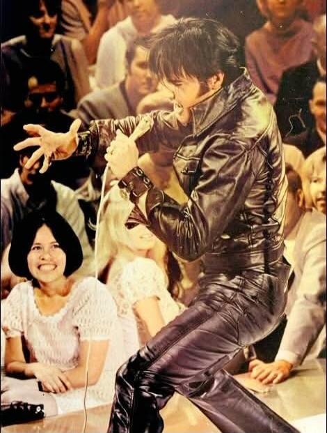 A man in a black leather suit is performing on stage, holding a microphone and interacting with the audience. A woman in the foreground is smiling widely.