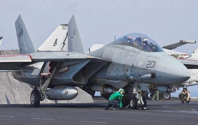 A military jet on a carrier deck with crew in green shirts around the aircraft. Military aircraft resembling an F/A-18 with the number 201 on the nose and 50 on the tail.