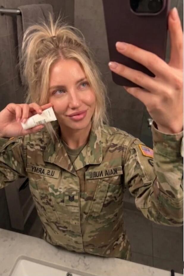 A blonde woman in a US Army uniform is taking a selfie in a bathroom mirror, holding a small white tube of product near her face. The uniform has 'U.S. ARMY' and an American flag patch visible (text is mirrored).