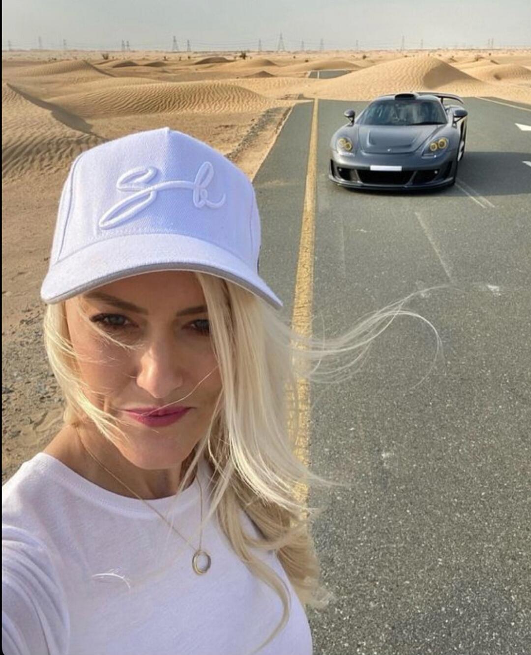A blonde woman in a white cap and t-shirt takes a selfie on a desert road. A grey sports car is parked behind her, with sand dunes visible in the background. The cap has embroidered initials 'gb'.
