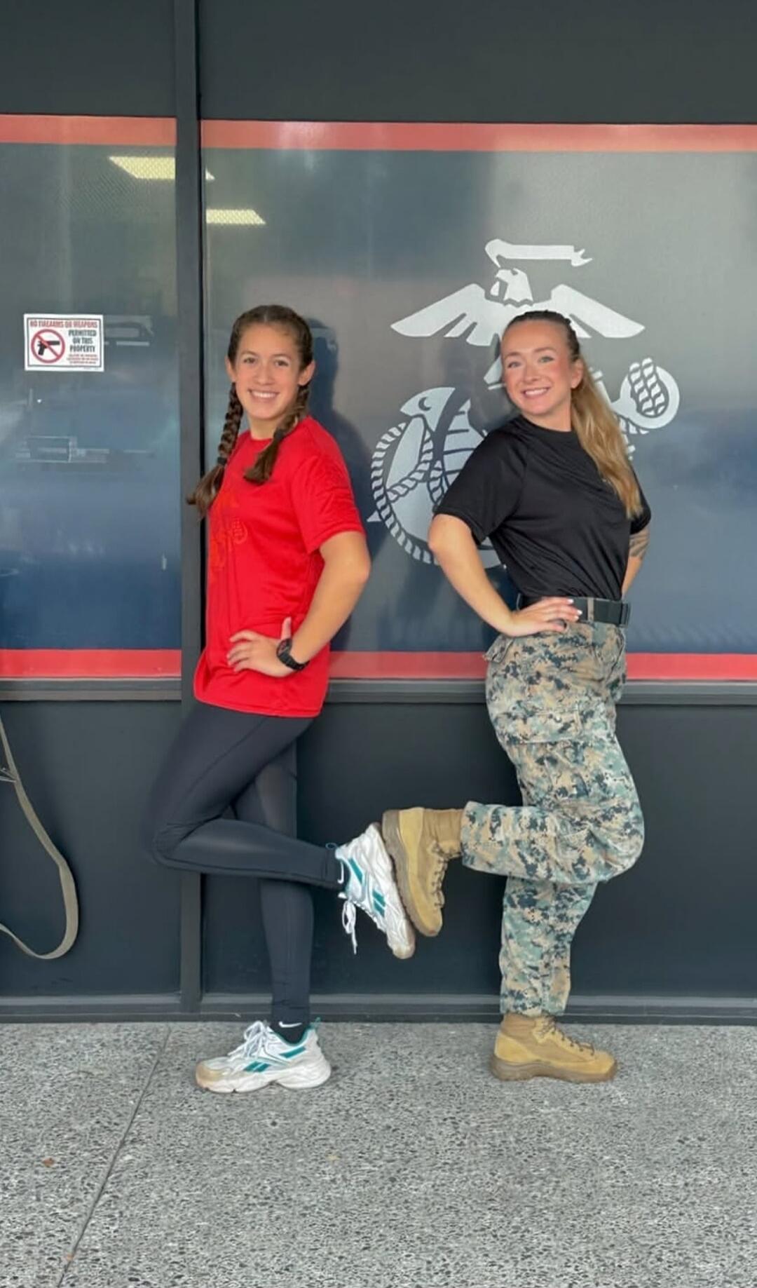 NO FIREARMS OR WEAPONS PERMITTED ON THIS PROPERTY. Two young women are posing in front of a wall with the US Marine Corps emblem. One woman is wearing a red t-shirt and black leggings, the other is in a black t-shirt and camouflage pants, possibly a military uniform. Both are smiling and have one leg bent.