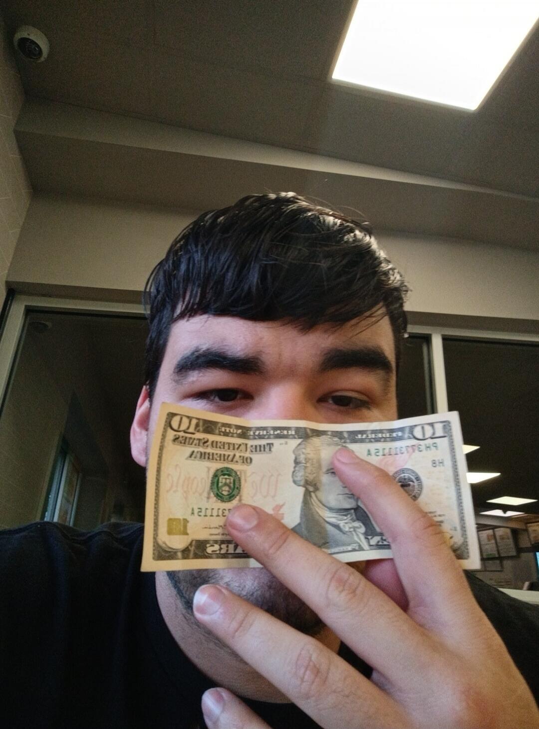 A person holding a 10 dollar bill, partially obscuring their face. The bill shows 'THE UNITED STATES OF AMERICA' and 'TEN DOLLARS' with Alexander Hamilton's portrait.