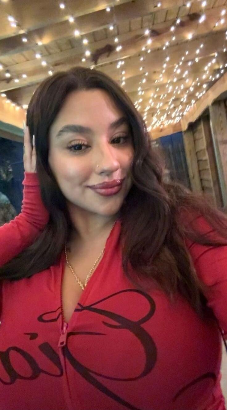 A woman with long dark hair wearing a red zip-up top with a black graphic, posing for a selfie under string lights.