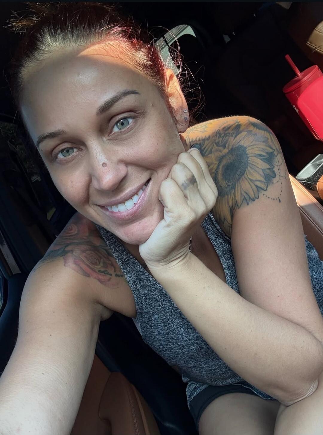 A woman with red hair and light eyes is smiling at the camera, resting her chin on her hand. She has tattoos on her shoulders, including a sunflower and roses. She is wearing a grey tank top and appears to be inside a car.