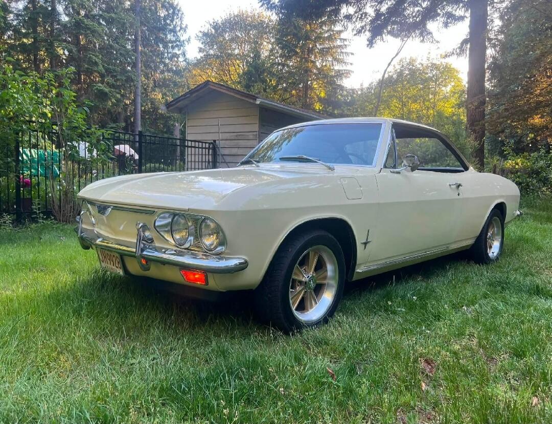 JH 9928. A vintage cream-colored car, possibly a Chevrolet Corvair, is parked on a lush green lawn. The background features tall trees and a small wooden shed under a bright sky.