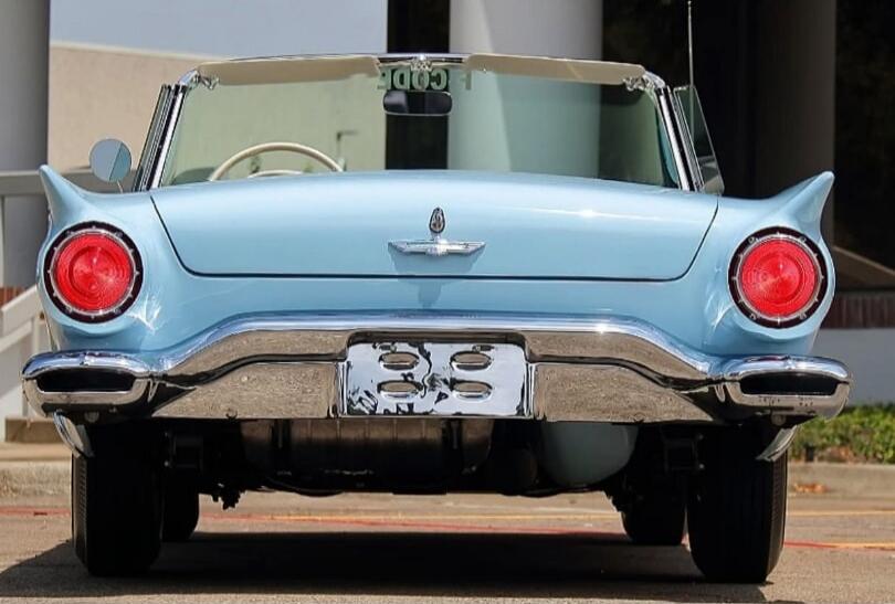 A rear view of a light blue vintage convertible car, possibly a Ford Thunderbird, with prominent round red taillights and chrome bumper. The text 'CODE' is visible in the rearview mirror.