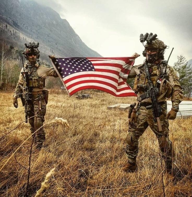 Two soldiers in camouflage holding a US flag in a field with mountains in the background.