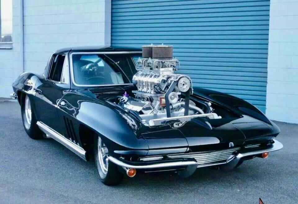 A black classic car with a large exposed engine on a driveway in front of a blue roll-up door.