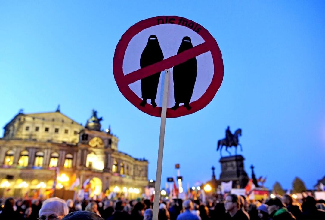A protest sign with a red circle and a diagonal line over two figures in burqas, with the text 'nie mals' (never). The sign is held amidst a crowd of people at a demonstration.