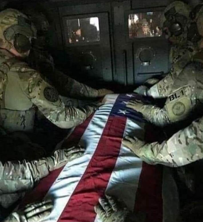 No visible text; image shows soldiers carrying a flag-draped coffin inside a military vehicle.