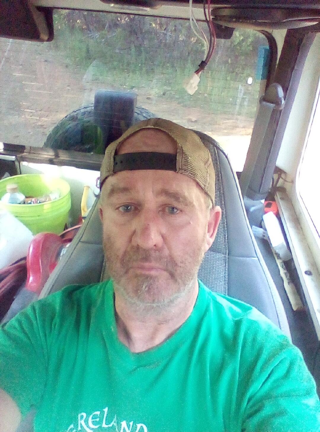 A man wearing a green t-shirt with 'RELAND' visible on it and a cap, sitting in a vehicle. He has a beard and is looking at the camera.
