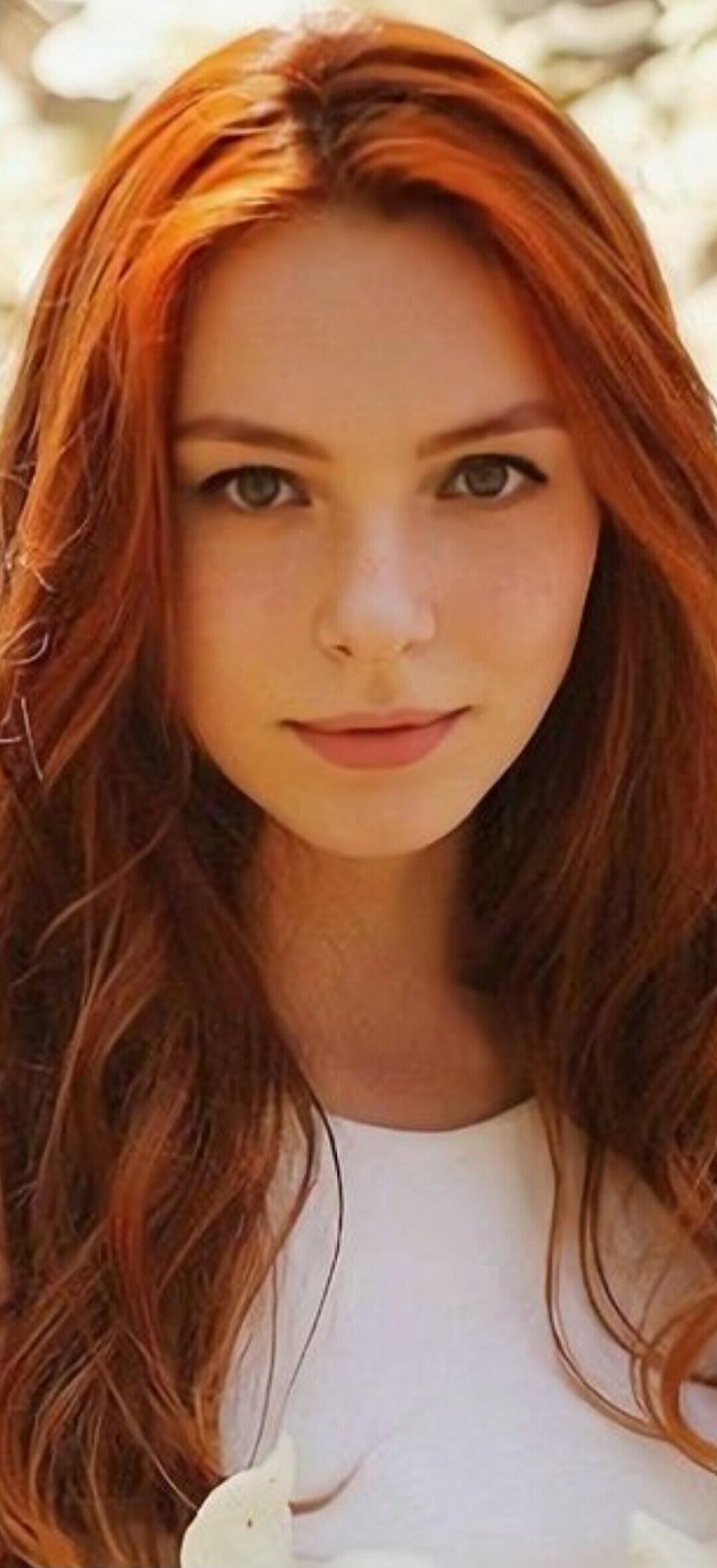 Portrait of a young woman with long red hair wearing a white top, outdoors.