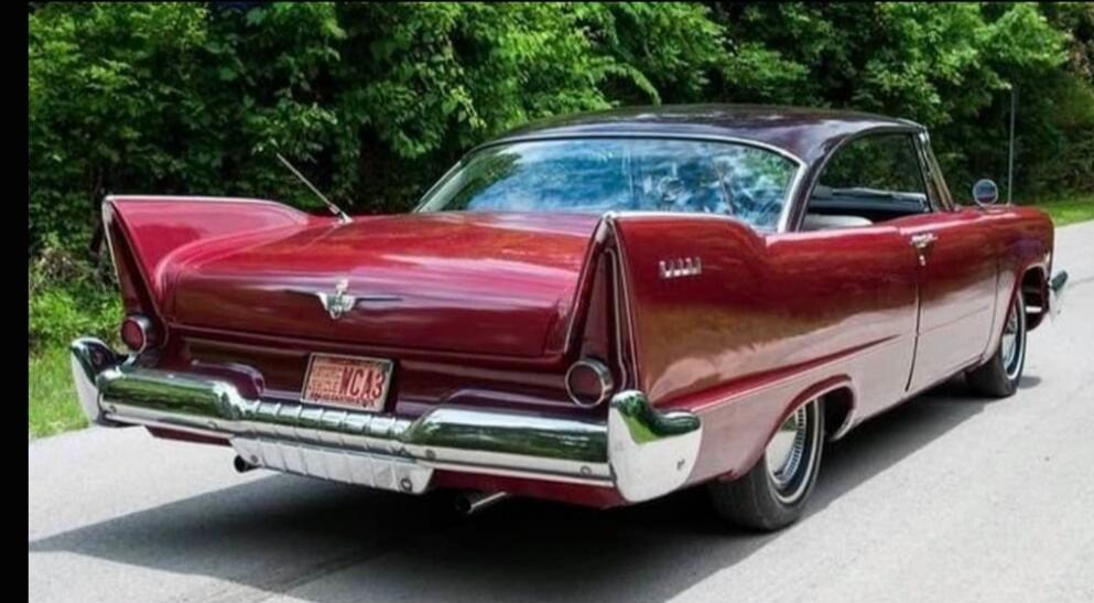 A vintage red car with prominent tail fins and chrome details, parked on a road with green trees in the background. The car has a dark roof and a shiny red body.
