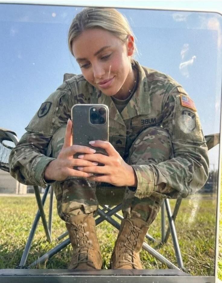 A young woman in a US Army uniform takes a selfie in a mirror. She is smiling and holding a smartphone. She is wearing combat boots and has manicured nails. The setting appears to be outdoors.