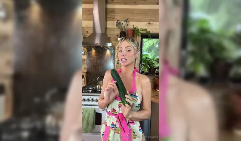 A woman in a revealing apron holds a large cucumber in a kitchen.