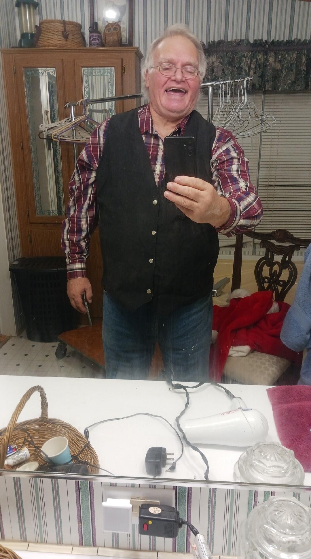 The man is taking a selfie in the mirror. He is wearing a plaid shirt, a vest, and jeans. He is laughing and holding a phone in his hand. He is also holding a knife in his other hand.