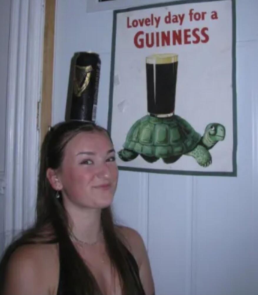 A woman is balancing a can of Guinness beer on her head, smiling at the camera. Behind her, a poster reads 'Lovely day for a GUINNESS' and depicts a turtle with a glass of Guinness on its shell.