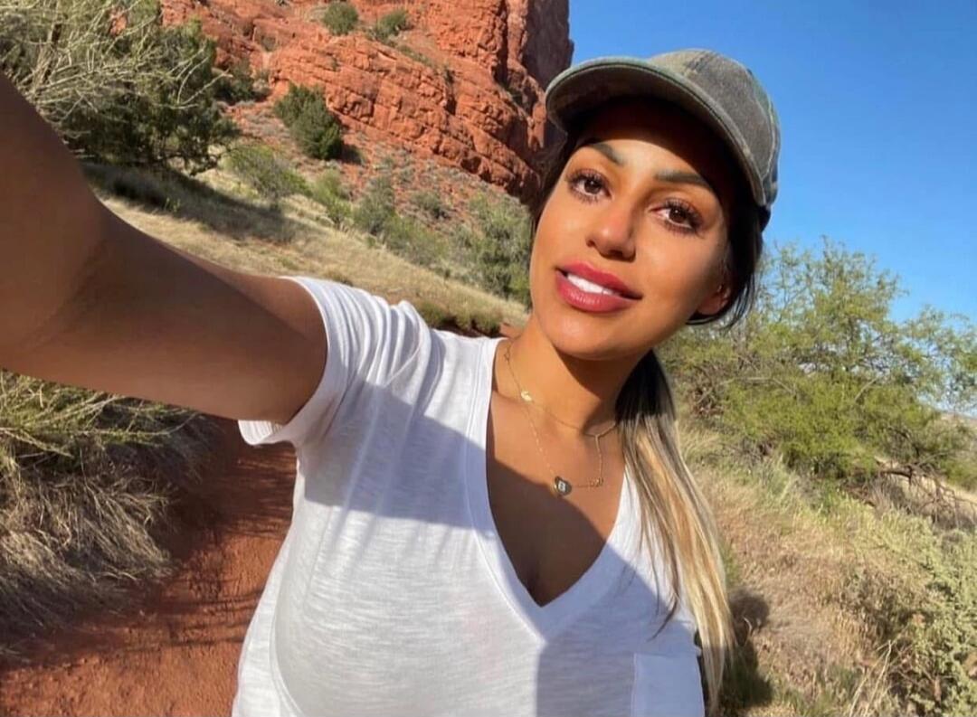 A woman wearing a white V-neck shirt and a gray cap taking a selfie outdoors, with red rock formations and desert vegetation in the background.