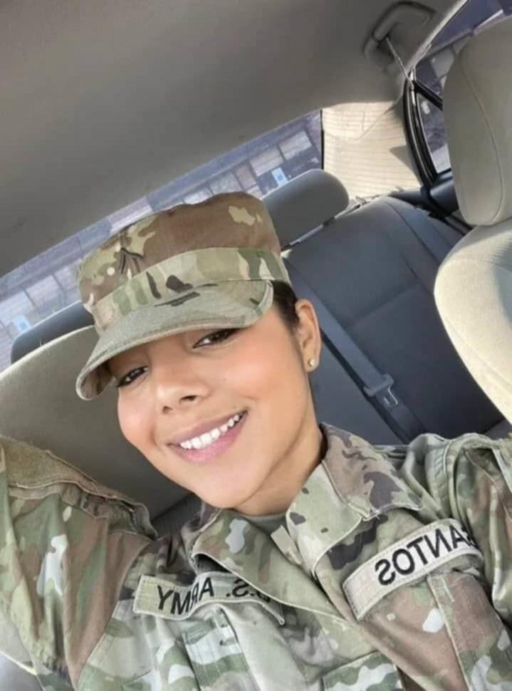 A smiling woman in a military uniform and cap takes a selfie inside a car. Text on uniform patches reads 'ARMY' and 'SANTOS'.