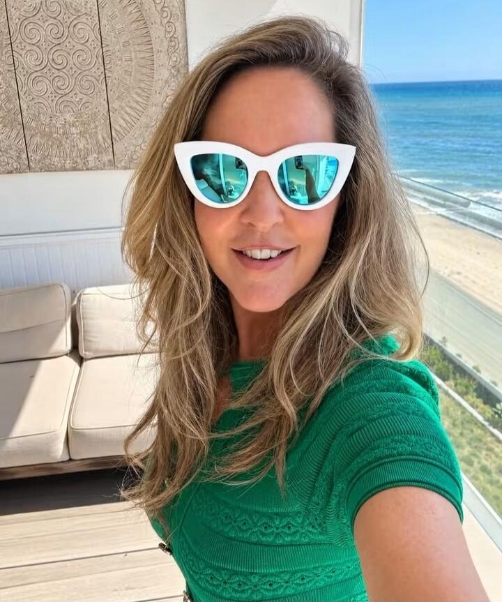 A woman takes a selfie on a balcony overlooking the ocean. She is smiling and wearing white cat-eye sunglasses with blue reflective lenses and a green top. The background shows a beach and the sea under a clear sky.