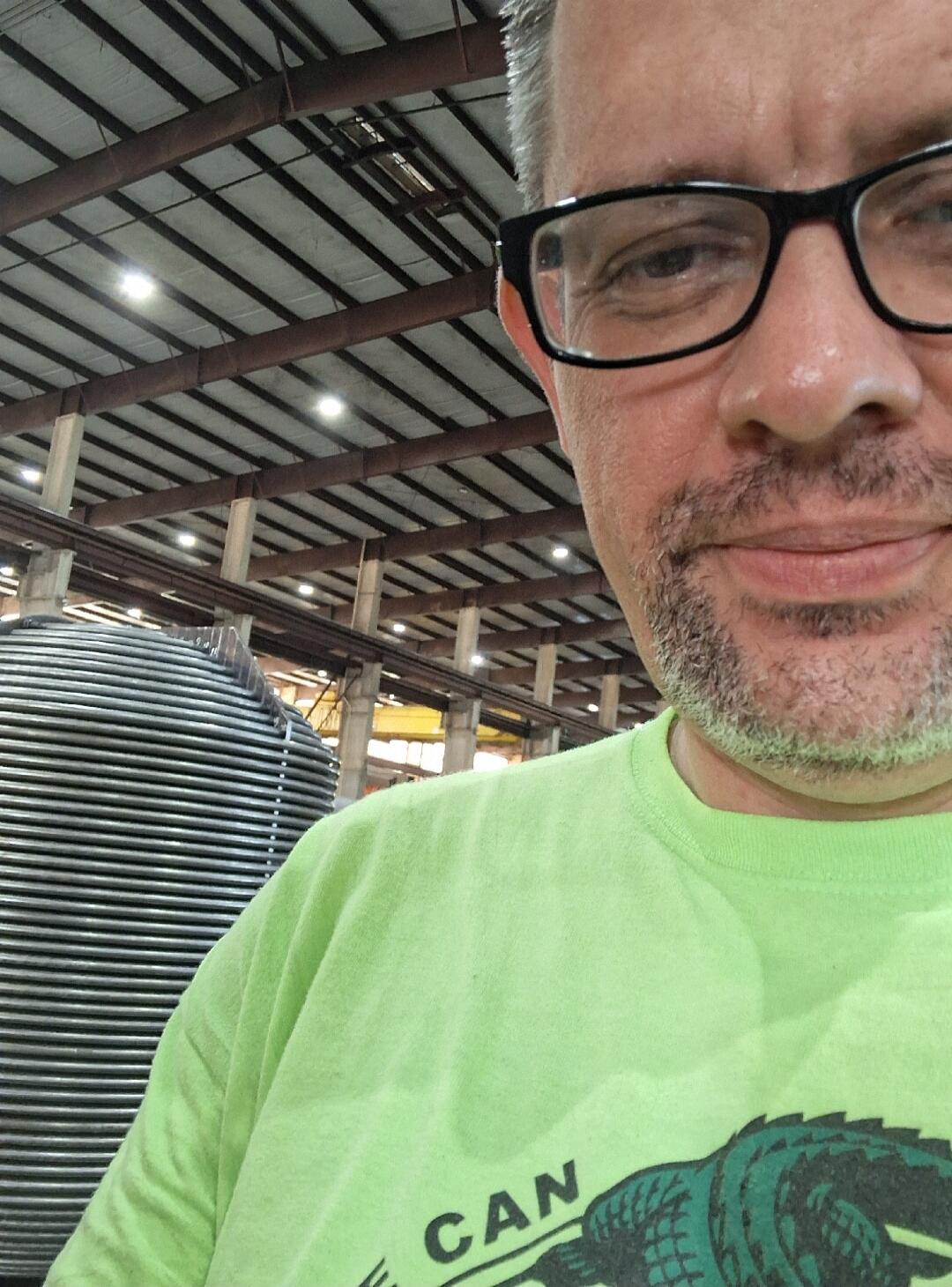 A man in glasses and a lime green t-shirt with a partial text 'CAN' and an alligator graphic, taking a selfie in what appears to be an industrial facility with a large metal structure in the background.