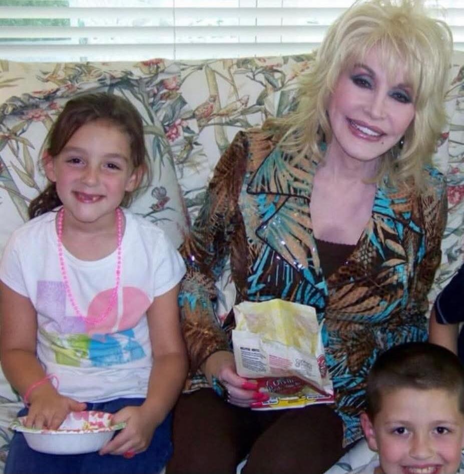 A smiling Dolly Parton sits on a couch with two children, a girl and a boy. Dolly is holding a snack bag, and the girl is holding a bowl.