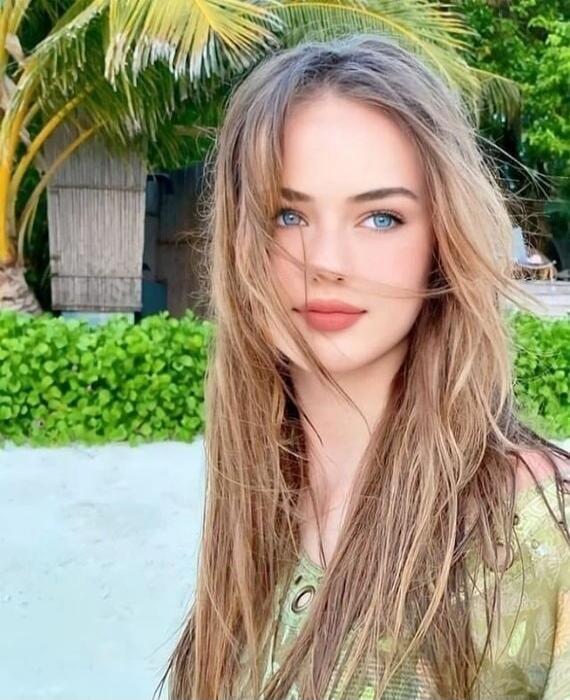 A young woman with long blonde hair, blue eyes, wearing a light green top, outdoors with palm trees and greenery in the background.