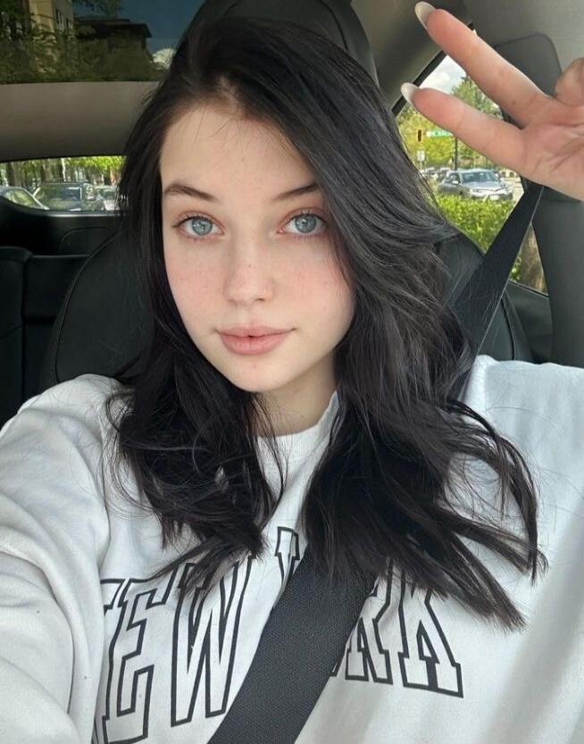 Selfie of a young woman in a car wearing a seatbelt and a white sweatshirt, flashing a peace sign.
