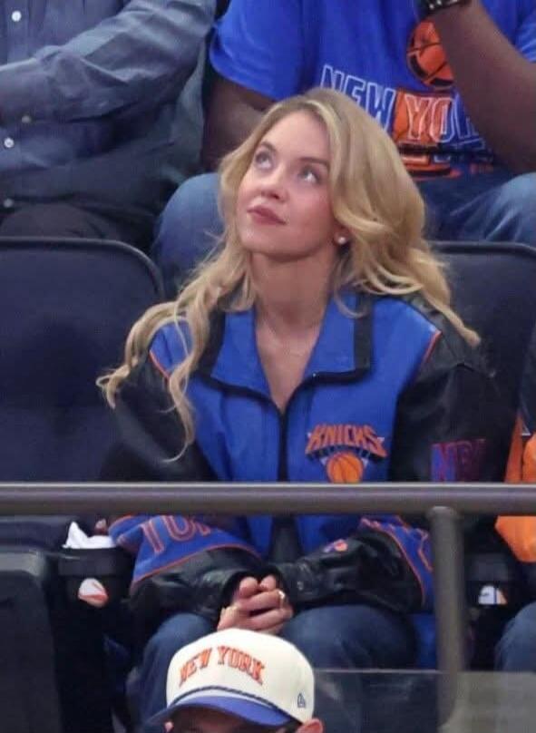 A blonde woman wearing a blue and black jacket with 'KNICKS' and a basketball logo, looking up. In the background, other spectators are visible, one wearing a 'NEW YORK' shirt. A person in the foreground wears a 'NEW YORK' cap.