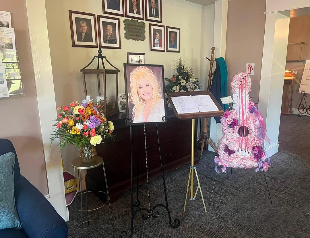 A room with a large portrait of Dolly Parton on an easel. To its left, a vibrant bouquet of flowers. To its right, a music stand with papers and a large floral arrangement shaped like a pink guitar, decorated with ribbons and butterflies. Several framed pictures hang on the wall in the background.