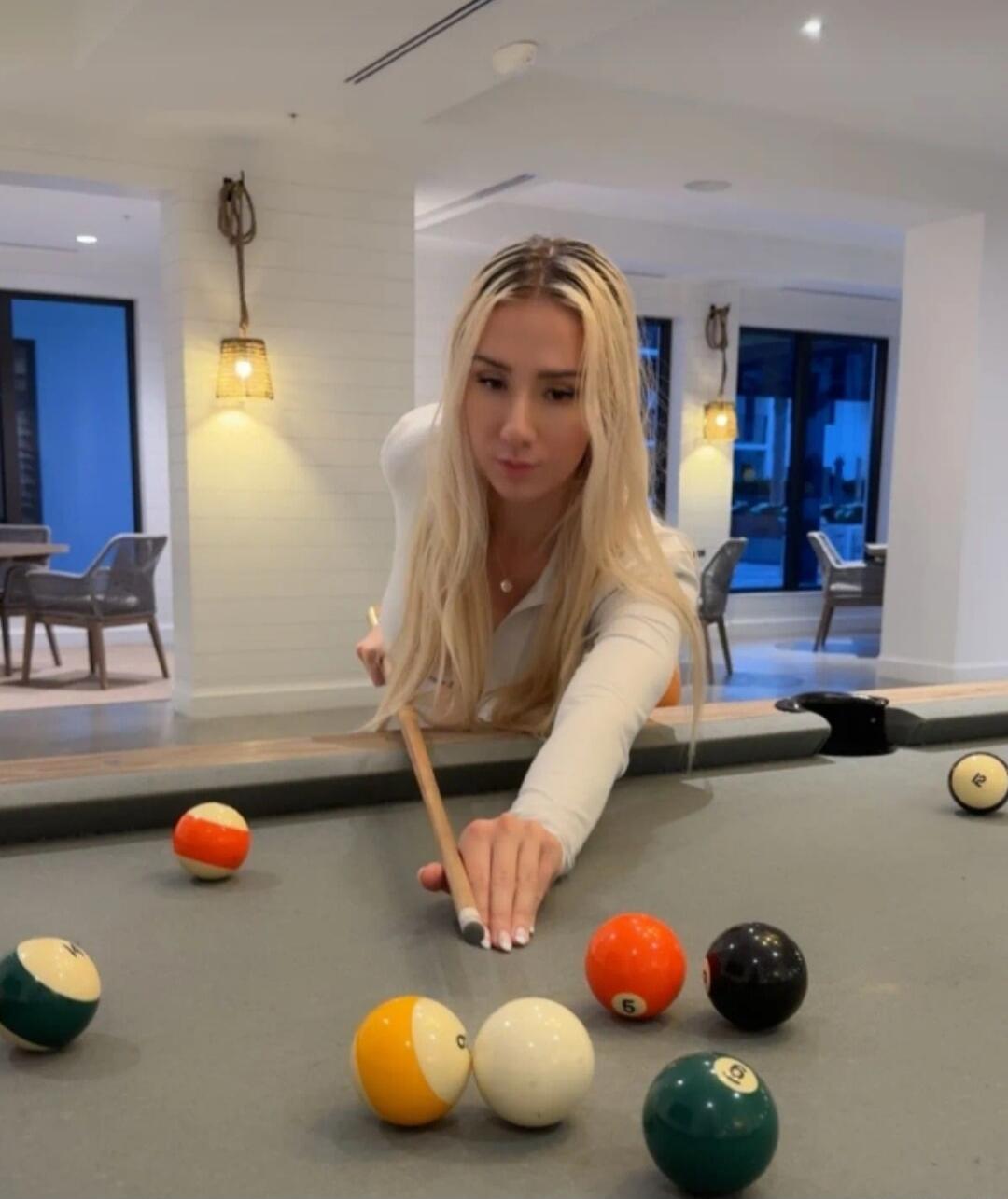 A blonde woman is playing pool, aiming with a cue stick at the balls on the table in a bright, modern room.