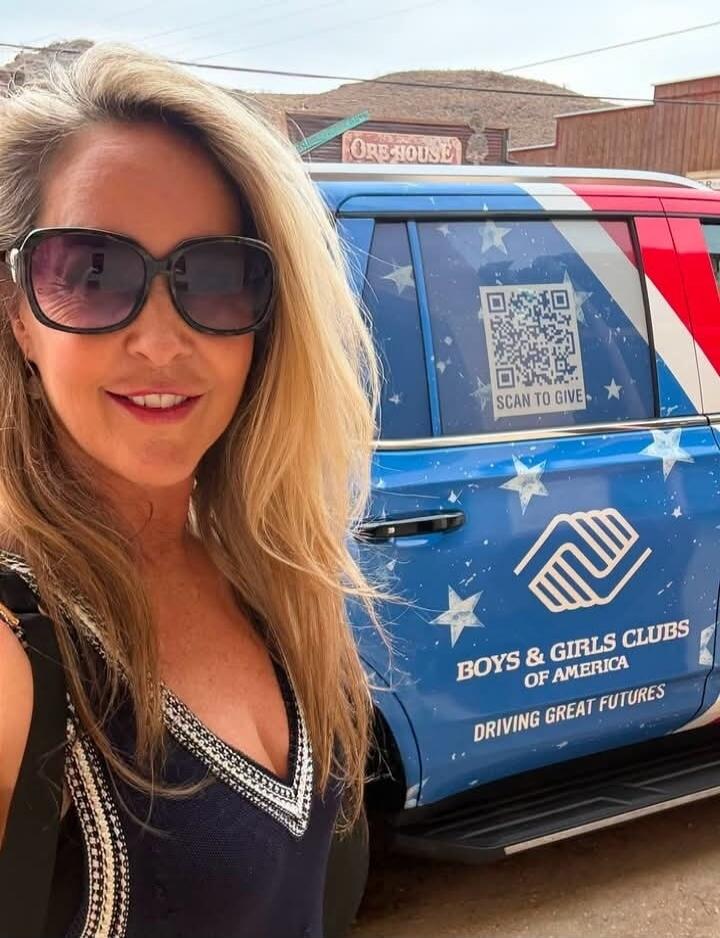 A woman in sunglasses takes a selfie in front of a blue vehicle. The vehicle is decorated with stars and stripes, featuring the logo and text 'BOYS & GIRLS CLUBS OF AMERICA' and 'DRIVING GREAT FUTURES'. A QR code with 'SCAN TO GIVE' is visible on the window. In the background, a building with an 'ORE HOUSE' sign can be seen.