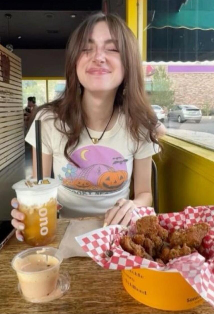 SPOOKY SEAS (on shirt), ono (on cup). A young woman is smiling with her eyes closed, enjoying a meal and a drink at a cafe.
