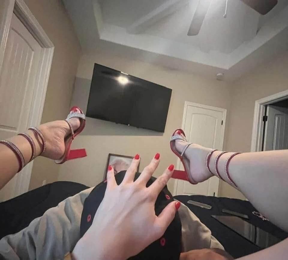 A person lying on a bed with their legs up in the air, wearing red high-heeled sandals and red nail polish. One hand covers part of their face.