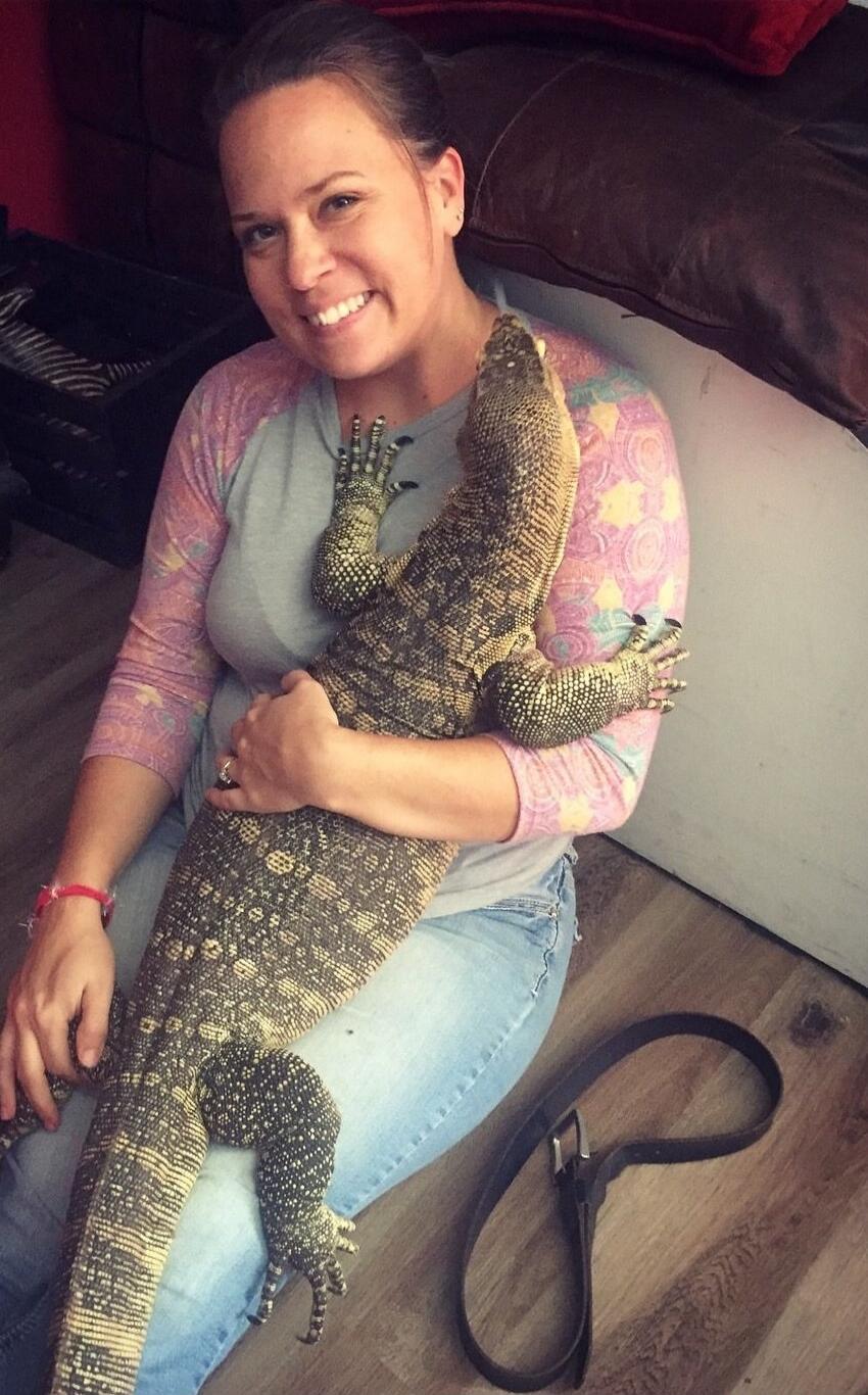 A woman sitting indoors on a chair/couch, smiling, while hugging a large alligator that is wrapped around her torso and arm.