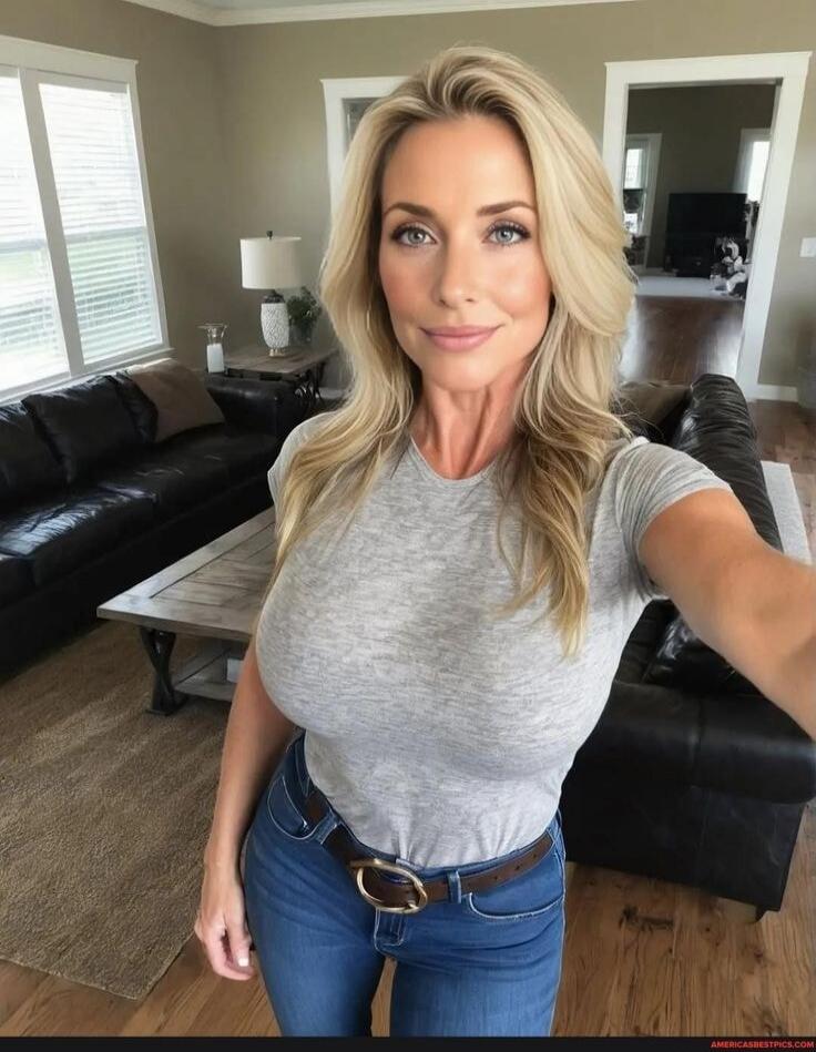 A woman with long blonde hair wearing a gray t-shirt and blue jeans takes a selfie in a living room with a black sectional sofa and a coffee table in the background.
