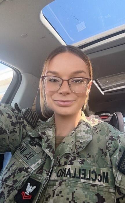 A young woman in a US Navy uniform, wearing glasses, smiles at the camera while taking a selfie in a car. The name 'MCCLELLAND' and 'U.S. NAVY' are visible on her uniform.